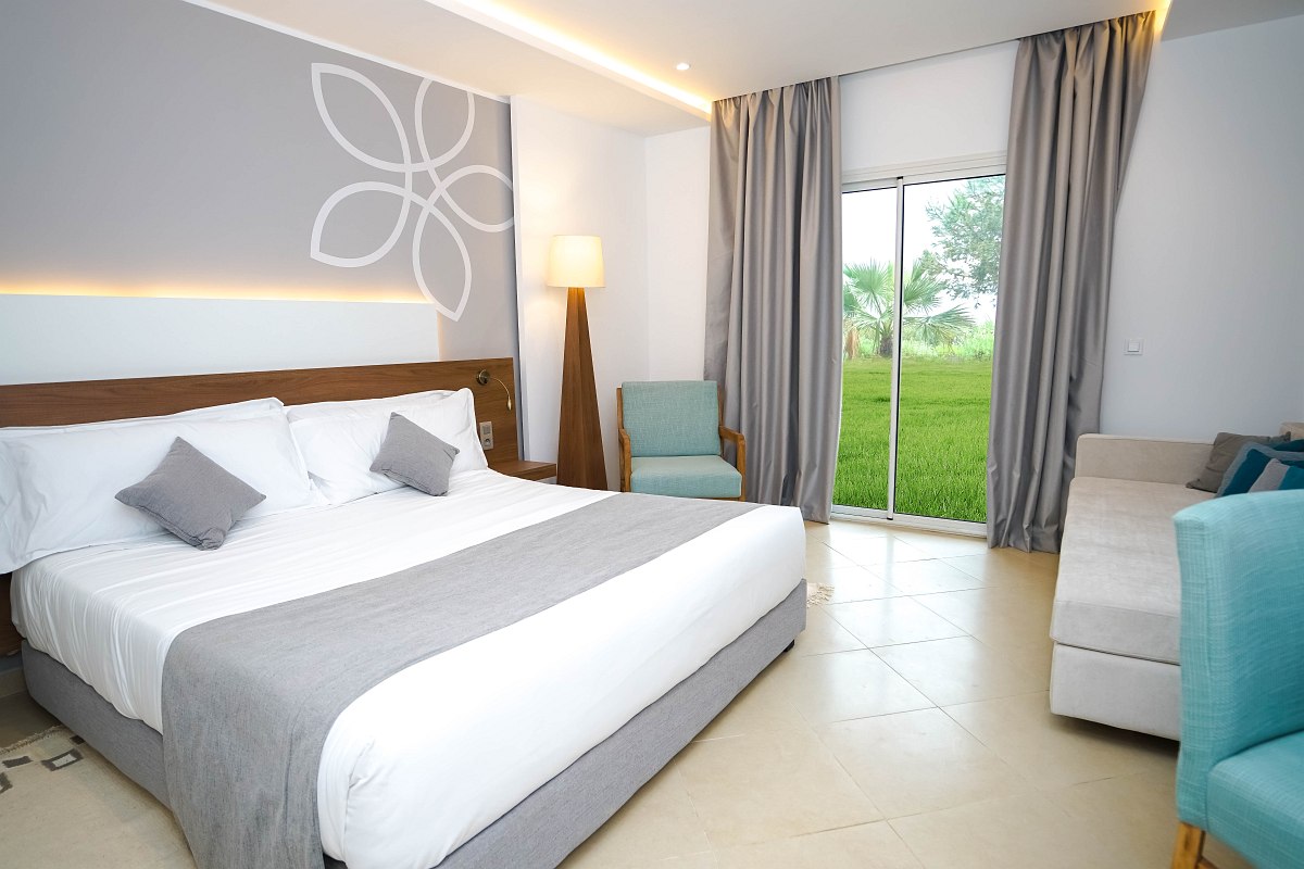 Brightly furnished Hotelroom with modern double bed, light blue armchair and kitchenette with coffee machine.