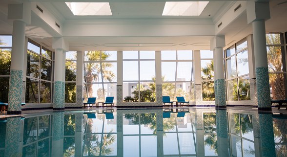 Brightly designed hotel lobby area with high windows, pillars with blue mosaic and palm tree view in the background.
