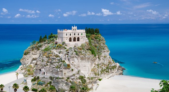 Drone view of rocky island of Tropea with the white church of Santa Maria dell'Isola on it, surrounded by turquoise-coloured sea and white sandy beach under a blue sky.
