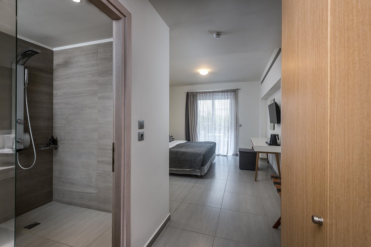 View from the entrance into a modern hotel room with double bed, balcony, desk and open-plan bathroom with walk-in shower.