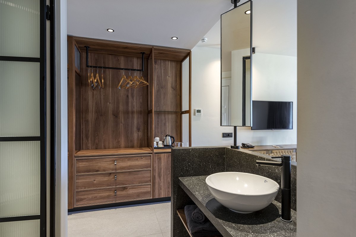 Open-plan hotel bathroom with modern washbasin, countertop basin, large mirror and view of the walk-in wooden wardrobe with tea station.
