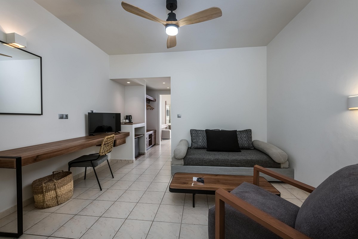 Bright living area with sofa, armchair, ceiling fan, desk, TV and view of the neighbouring kitchenette of a hotel room.