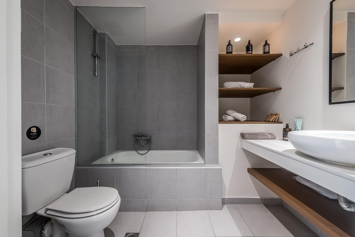 Modern bathroom with grey tiles, bathtub with shower screen, WC, washbasin and open shelves with towels.