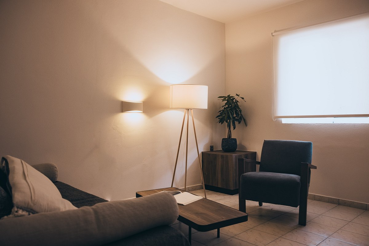 Cosy living area with armchair, sofa, floor lamp, small side table and plant in front of a darkened window.