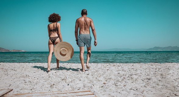 Two people in swimwear walk across a sandy beach towards the calm sea, with a clear sky in the background.
