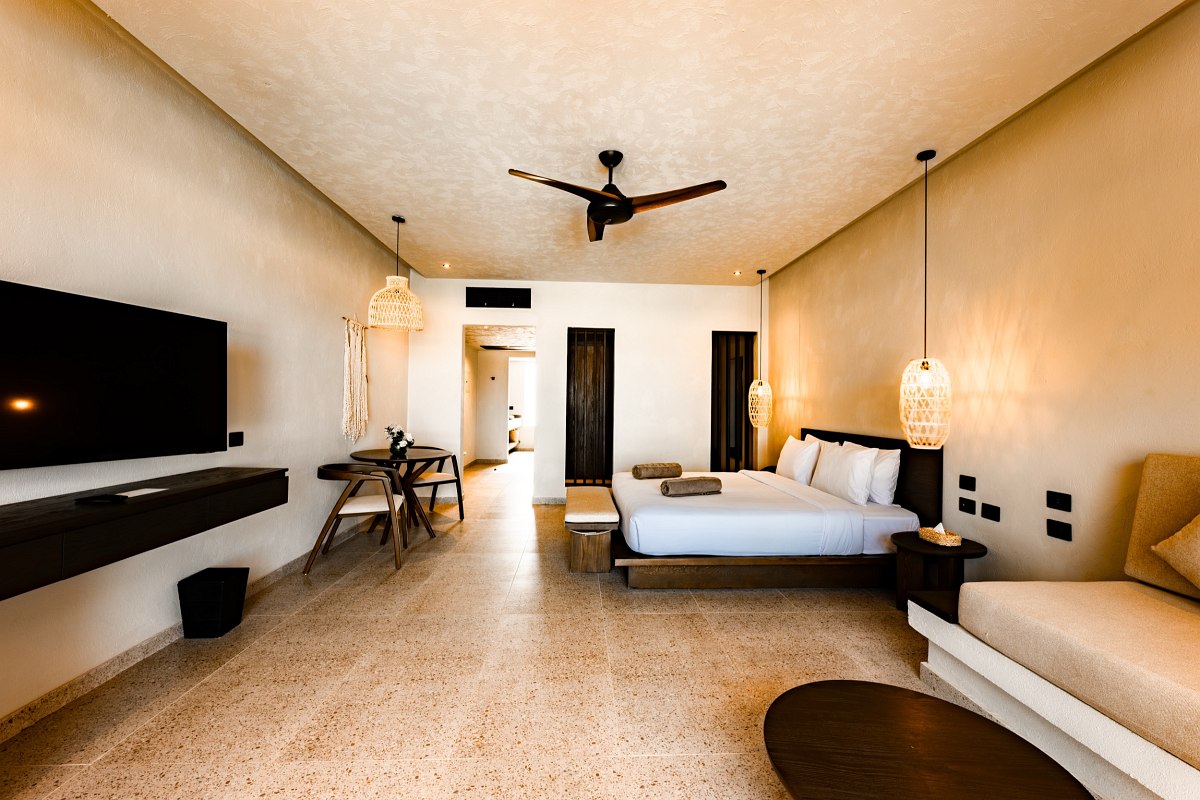 Modern hotel room with double bed, ceiling fan, seating area, TV and round mirror console.’