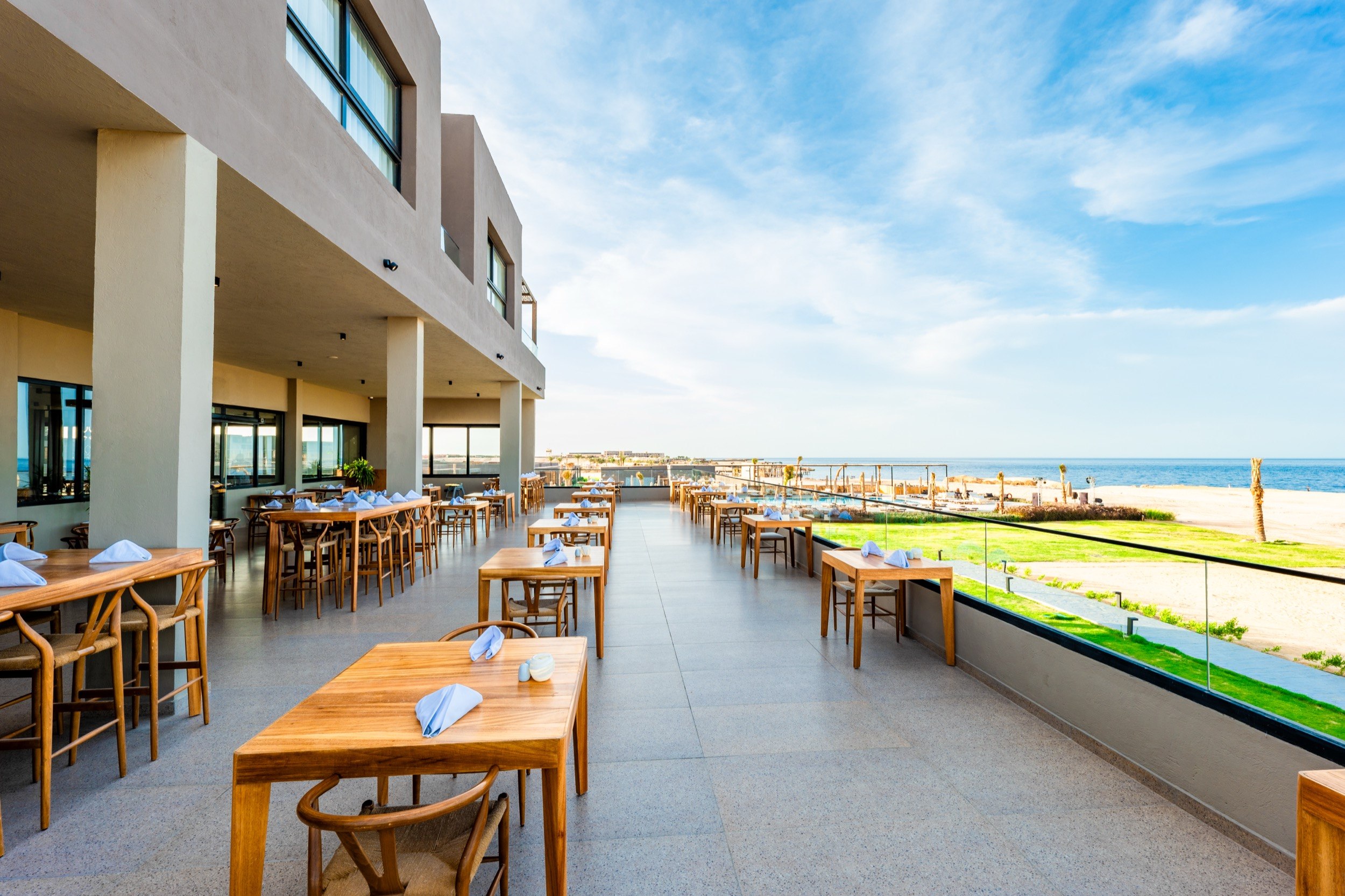 ‘Spacious restaurant terrace with wooden tables, view of the sandy beach and the sea under a blue sky.’