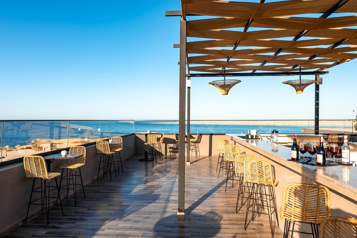 Roof terrace bar with rattan bar stools, sea view and pergola made of fabric panels under a blue sky.’