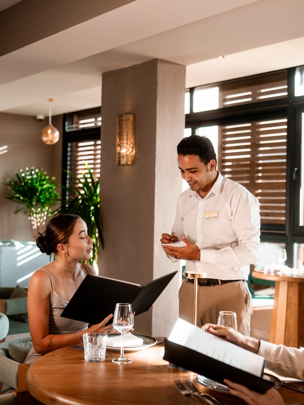 Restaurant scene with a waiter taking an order while guests sit at a table in a cosy atmosphere.