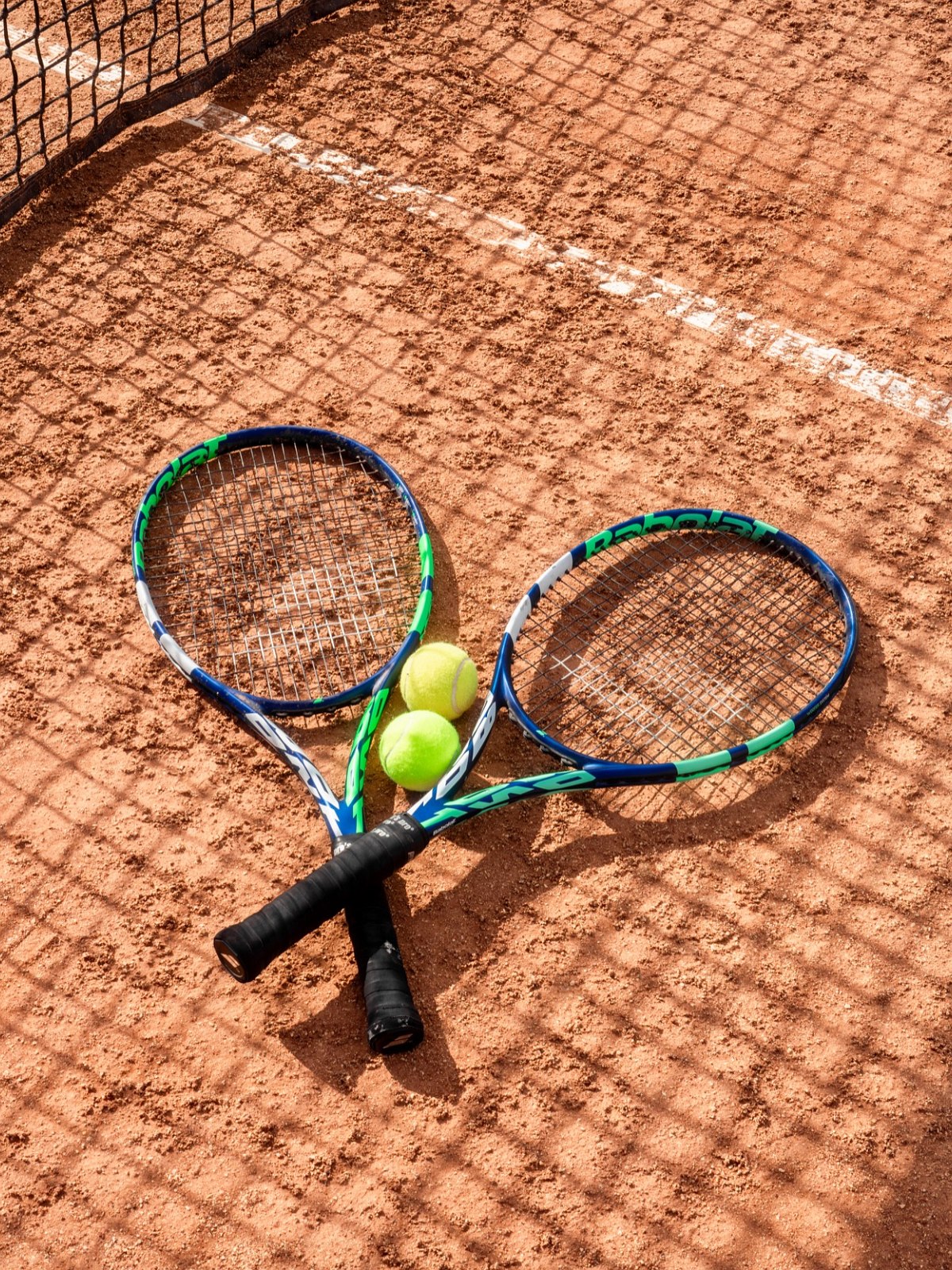 Two tennis rackets and three balls lie on a clay court under the shade of the net