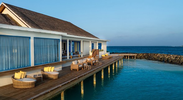  Terrace with wicker chairs and sofas on stilts over clear sea water, adjacent to a building with large windows and blue sky.