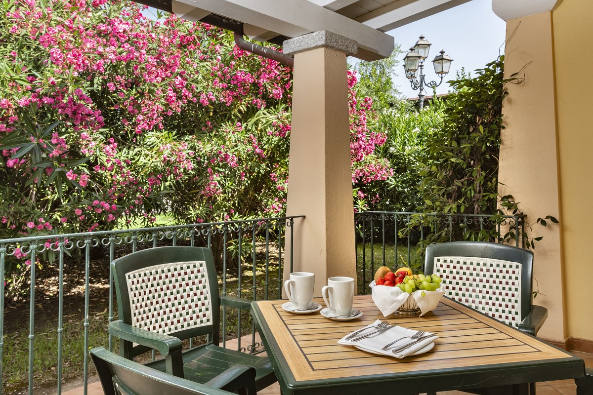 Covered terrace with garden table, chairs and breakfast dishes, surrounded by flowering oleander bushes.