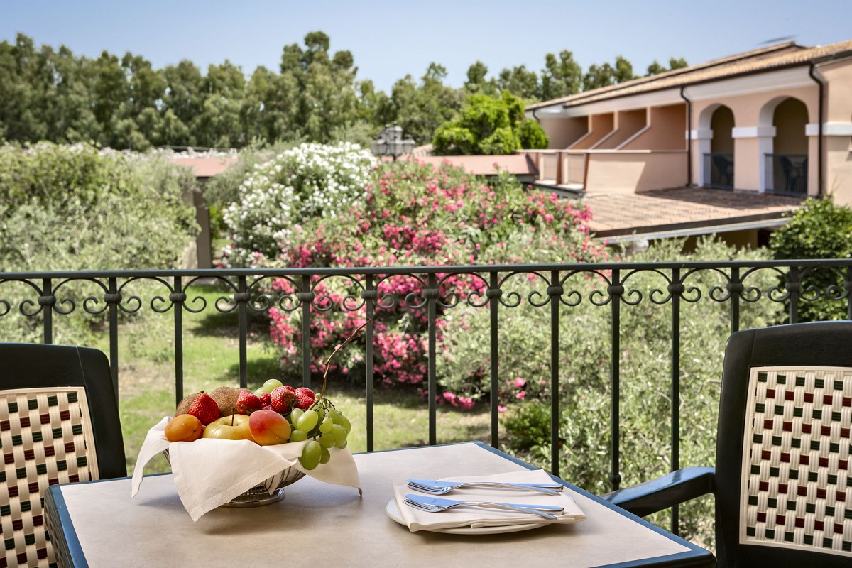 Balcony with seating area and fruit bowl, view of flowering garden and Mediterranean buildings in quiet, green surroundings.