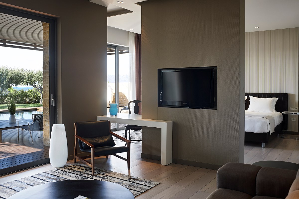 Modernly furnished hotel suite with living and sleeping area, desk, flat-screen TV and access to the terrace with pool view.