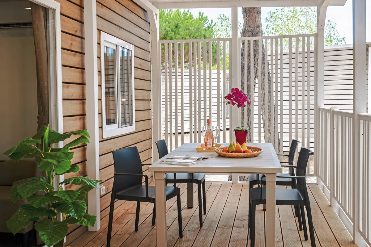  Modern mobile home with covered wooden veranda, table and four chairs, white railings and wide steps - surrounded by sandy terrain and pine trees.