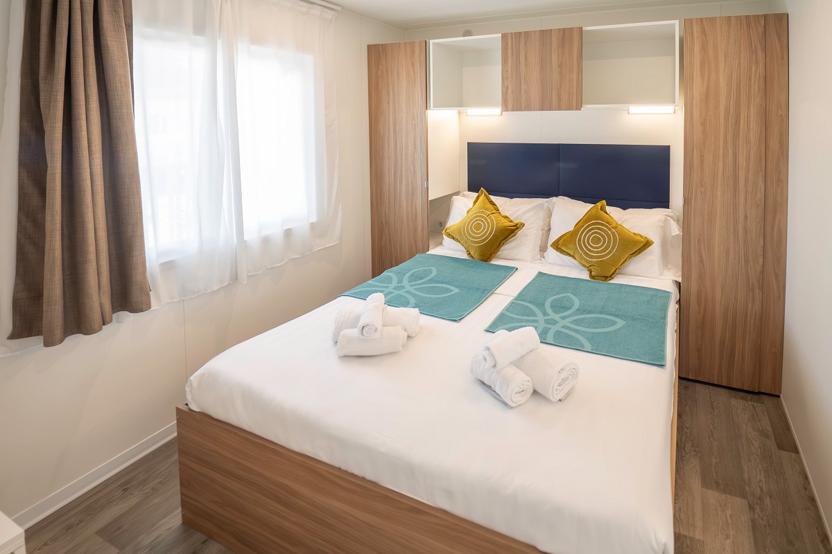 Modern bedroom in the mobile home with double bed, blue headboard, yellow cushions, wooden details and window - bright and cosy.