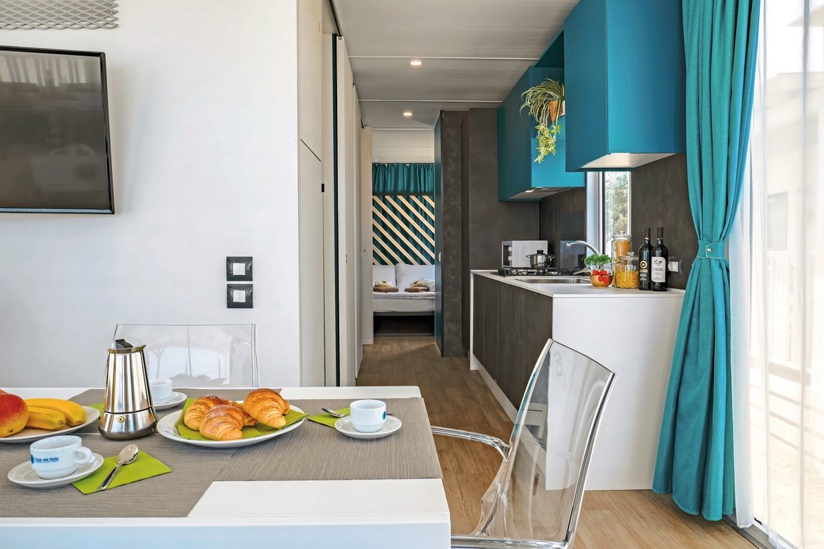 Camp Lodge Deluxe - Modern mobile home with open-plan kitchenette, dining table with breakfast, transparent chairs and view into the bedroom - bright and friendly.