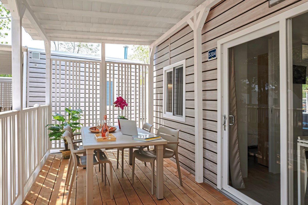  Modern mobile home with covered wooden veranda, table and four chairs, white railings and wide steps - surrounded by sandy terrain and pine trees.