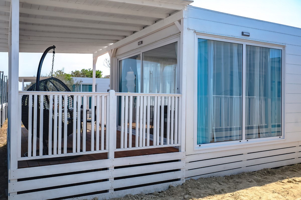 Modern mobile home with large covered wooden terrace, hanging chairs, white railings and floor-to-ceiling windows - inviting and bright.