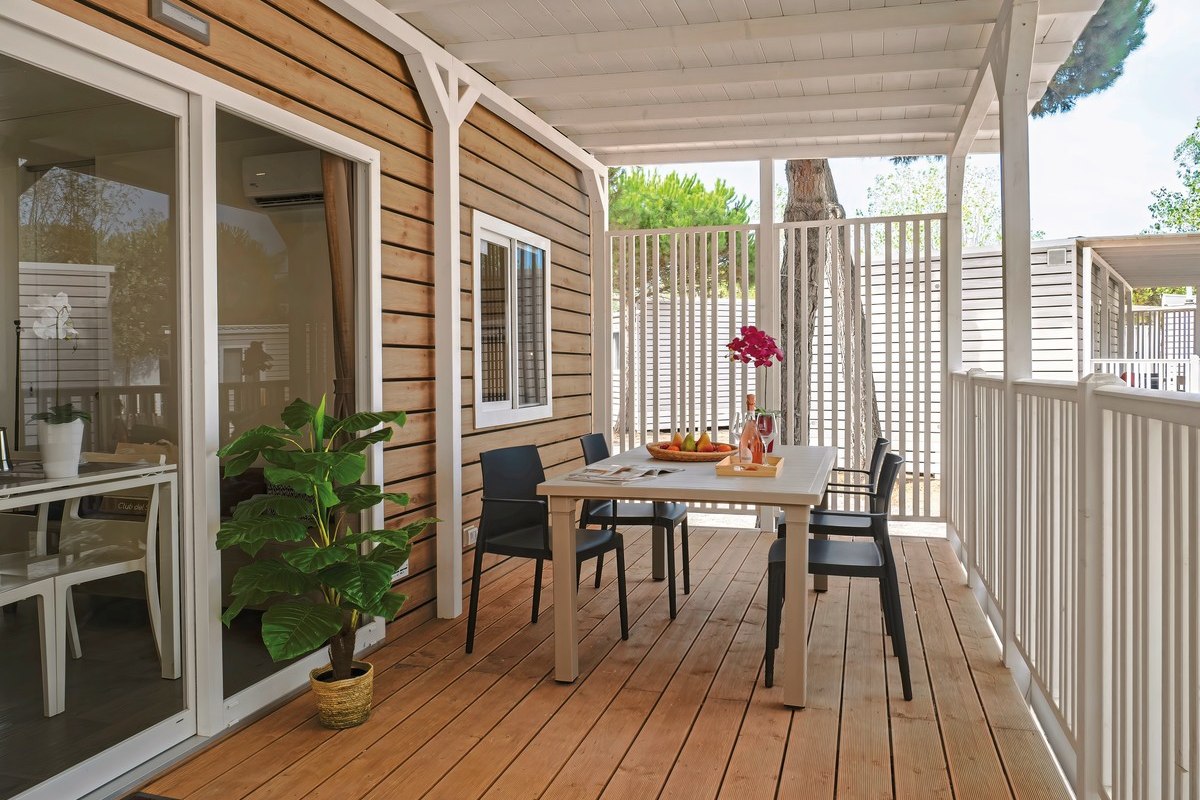  Modern mobile home with covered wooden veranda, table and four chairs, white railings and wide steps - surrounded by sandy terrain and pine trees.