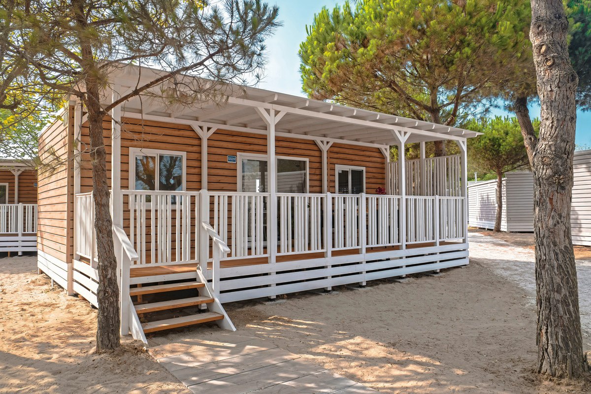 Camp Lodge Superior - Modern mobile home with covered wooden veranda, white railings and wide steps - surrounded by sandy terrain and pine trees.
