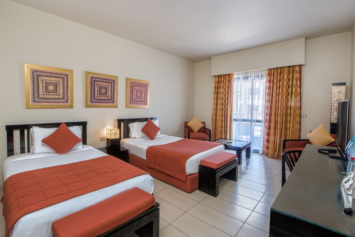 Cozy hotel room with two single beds, red bedspreads, warm lighting, a desk with TV, and orange curtains on the window.