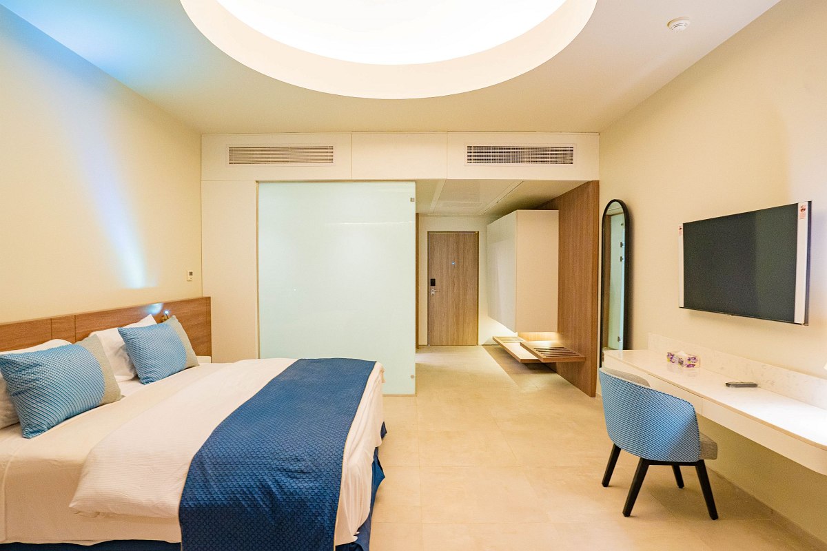 Modern, spacious room with beige walls and floors. Features a comfortable bed with blue accents, a desk with a matching chair, a wall-mounted TV, and a sliding glass door leading to a hallway. Bright natural light fills the room.