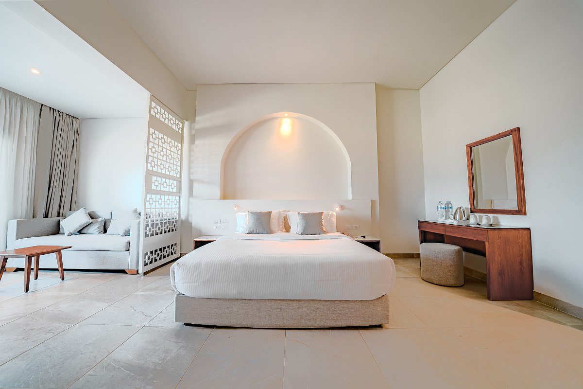 Bright, airy room with white walls and arched details. Features a blue-accented bed, tiled floor, and a seating area separated by decorative panels. Spacious and inviting.