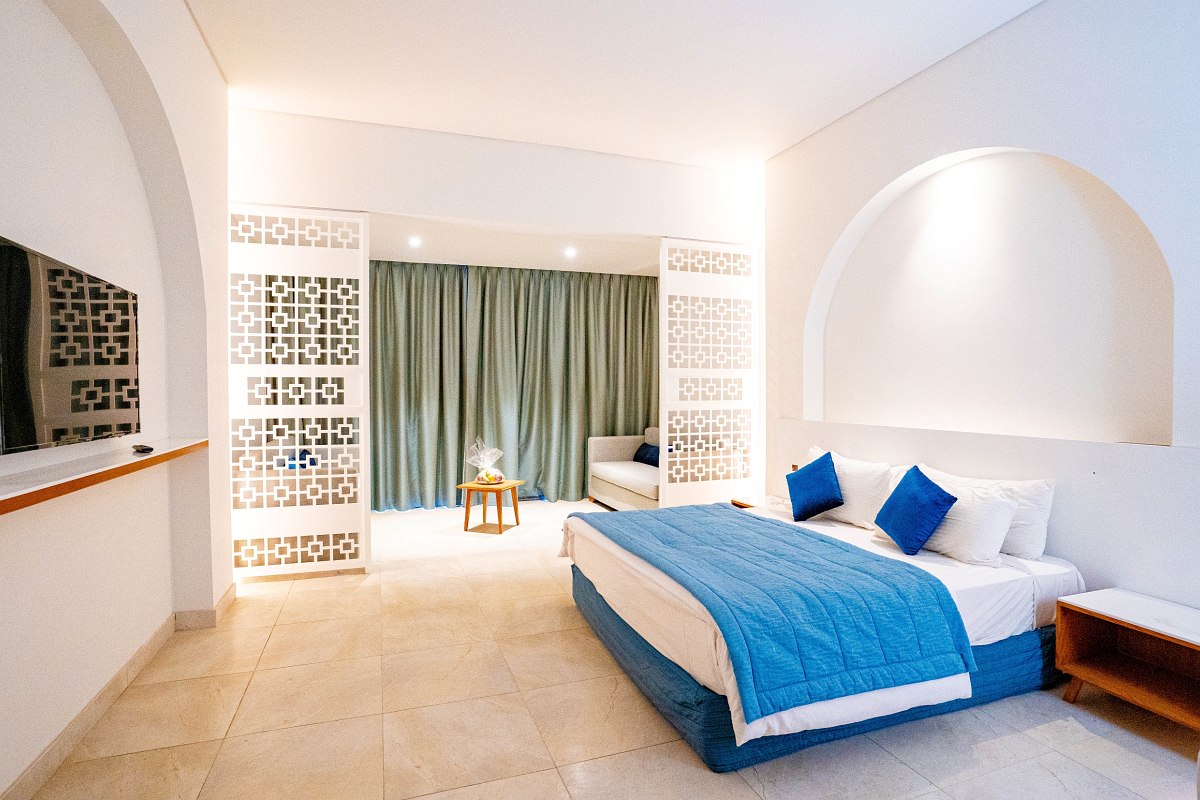 Bright, airy room with white walls and arched details. Features a blue-accented bed, tiled floor, and a seating area separated by decorative panels. Spacious and inviting.