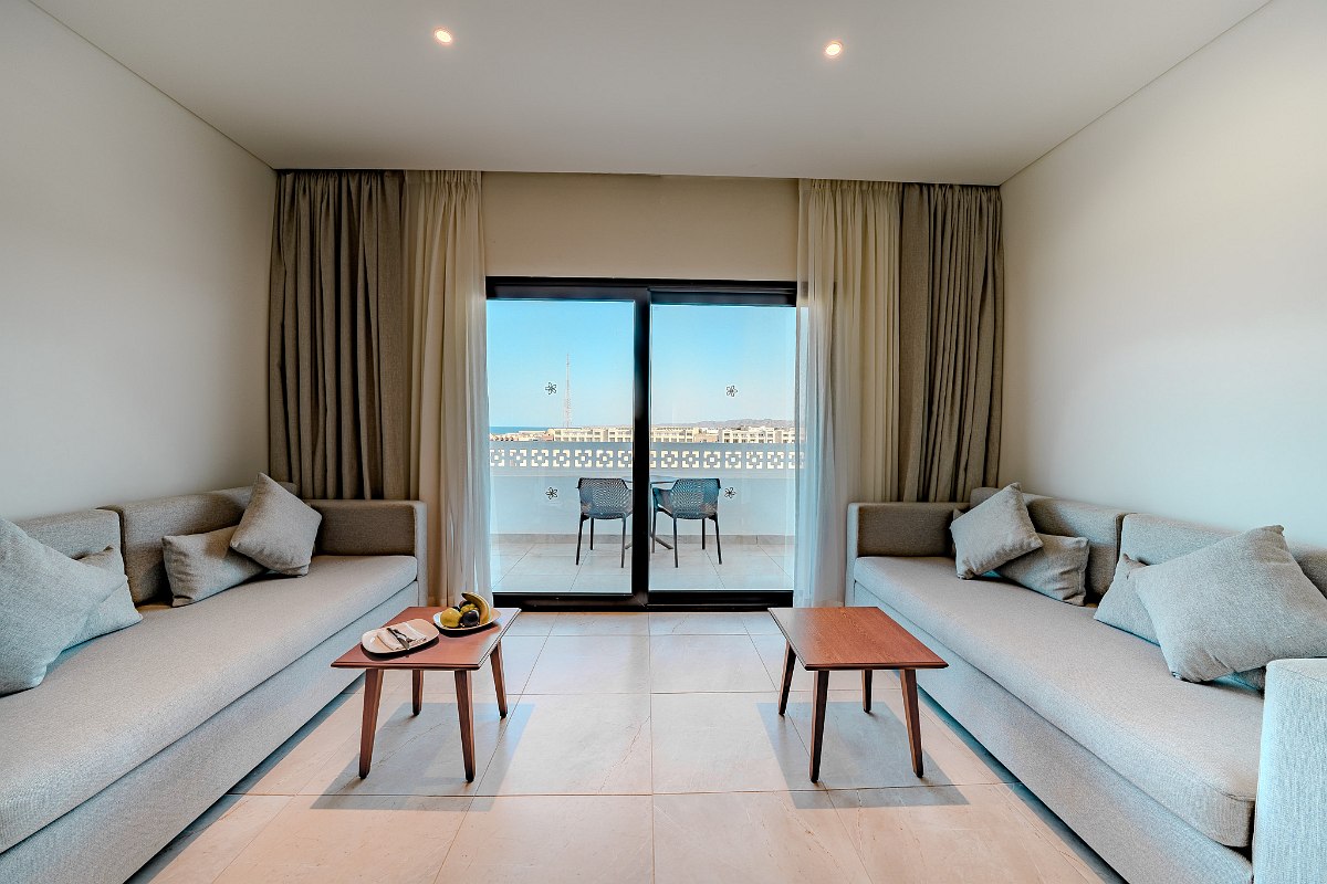 Sunny balcony with two wicker chairs and a view over white walls and rooftops, extending to the clear blue sea under a bright sky.