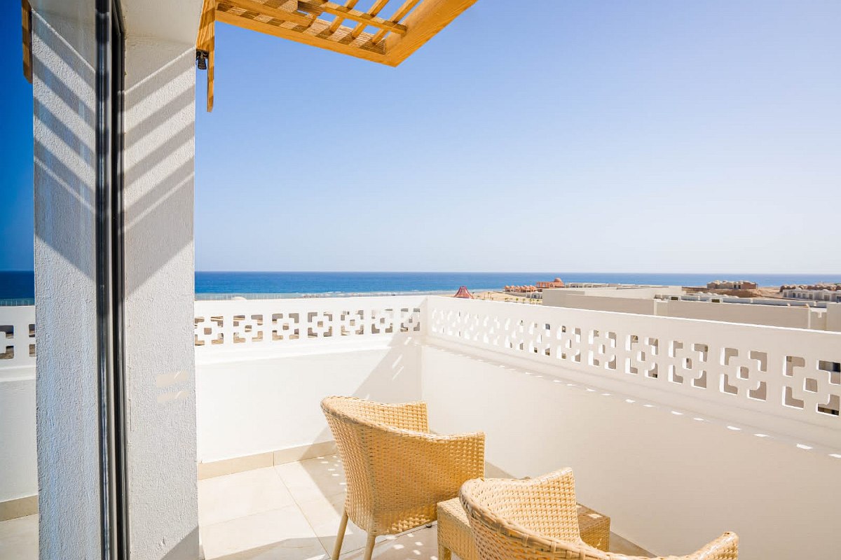 Sunny balcony with two wicker chairs and a view over white walls and rooftops, extending to the clear blue sea under a bright sky.