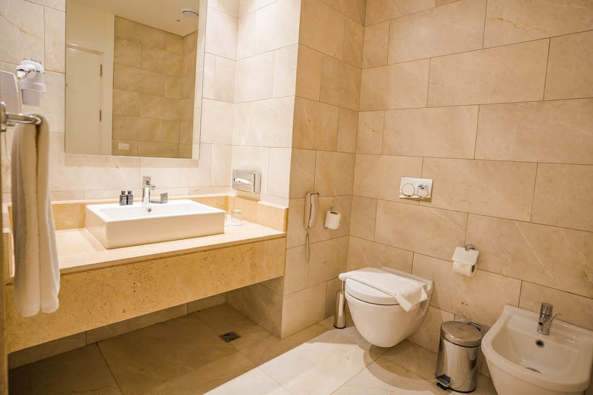 Clean, modern bathroom with beige tiled walls and floor. Features a wall-mounted toilet, bidet, large mirror, rectangular sink on a beige countertop, and towel rack.