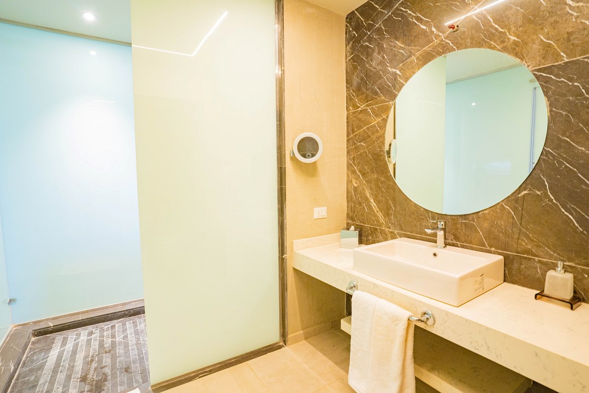 Modern bathroom with sleek beige tiles, a large circular mirror above a white rectangular sink, a floating countertop with a towel rack, and a frosted glass door leading to the shower area.