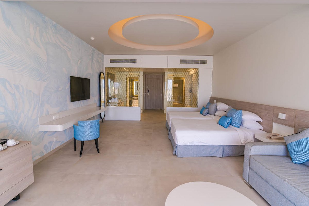 Bright and spacious hotel room with two beds adorned with blue pillows, a matching blue armchair, a sofa, a round white table, a wall-mounted TV, and a large mirror near the entrance. The room features light-colored floors, soft blue wallpaper with a subtle pattern, and a modern ceiling design.