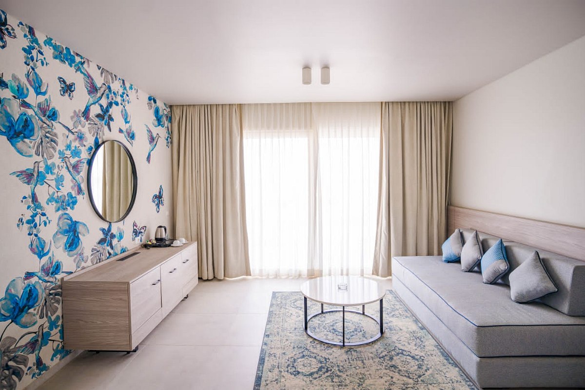 Modern and airy living area featuring a large window with sheer curtains, a gray sofa with five cushions, a round glass coffee table, a light patterned rug, and a wooden sideboard. The room has white walls, with one wall decorated in blue floral wallpaper, complemented by a round mirror.