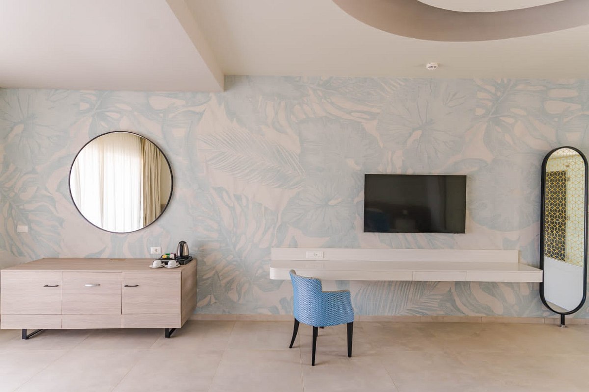 Bright room with floral wallpaper, round mirror, wooden dresser, floating desk, blue chair, TV, and tall oval mirror.