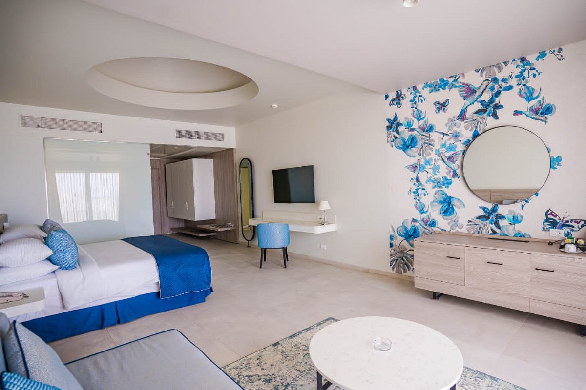 Spacious, light room with blue and white decor, floral wall art, large bed, sofa, round mirror, wooden dresser, desk, and TV.