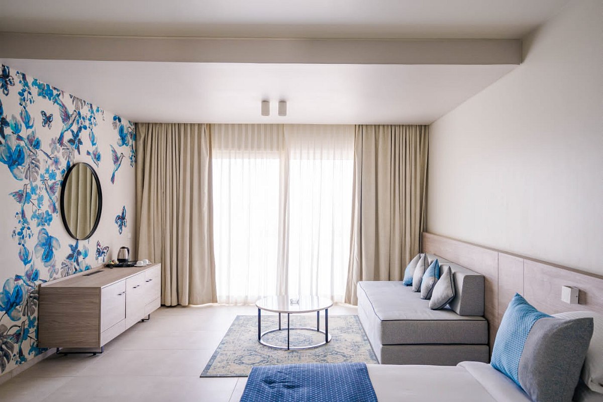 Modern and airy living area featuring a large window with sheer curtains, a gray sofa with five cushions, a round glass coffee table, a light patterned rug, and a wooden sideboard. The room has white walls, with one wall decorated in blue floral wallpaper, complemented by a round mirror.