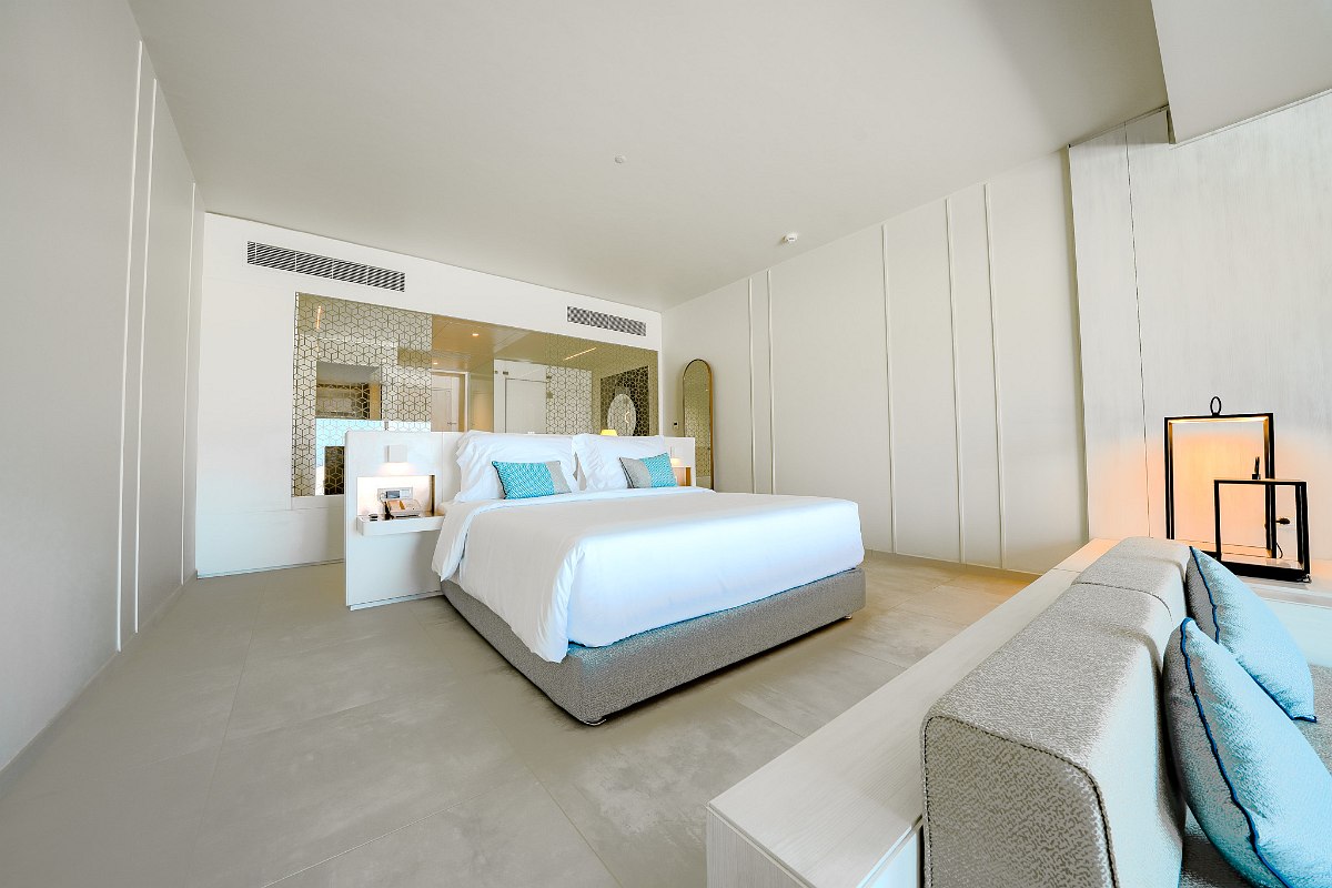 Modern hotel room with neutral tones, blue accents, TV, kettle, and large window overlooking the pool and ocean.