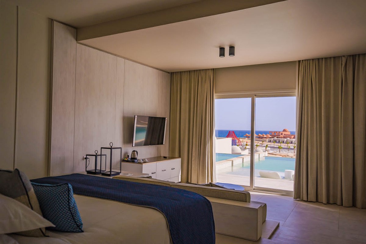 Modern hotel room with neutral tones, blue accents, TV, kettle, and large window overlooking the pool and ocean.