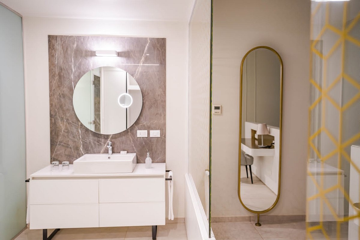 Modern bathroom with a white vanity, round mirror, marble wall, and a tall gold-framed mirror opposite the sink. Clean and minimalistic design.