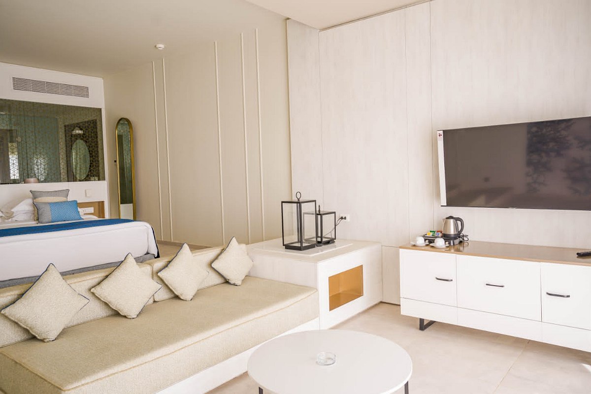 Bright, modern hotel room with a beige sofa adorned with matching cushions, a round white coffee table, and a white TV console with a flat-screen TV mounted on the wall. The bed is visible in the background with blue and white bedding, and a full-length mirror stands beside it.