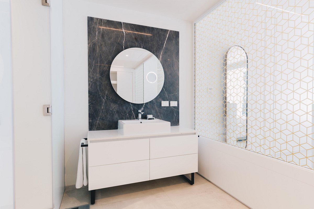 Bright, modern hotel room with a beige sofa adorned with matching cushions, a round white coffee table, and a white TV console with a flat-screen TV mounted on the wall. The bed is visible in the background with blue and white bedding, and a full-length mirror stands beside it.