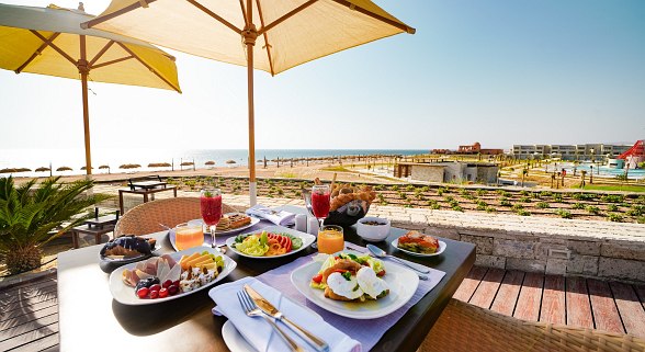 Breakfast table with a variety of food and drinks under parasols, with a view of the beach, sea and hotel grounds.