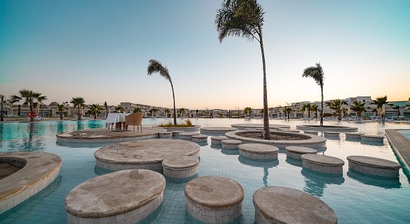Spacious pool area with round seating islands in the water, palm trees and a stylishly laid table at sunset.