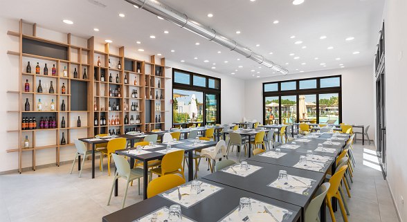 Brightly furnished dining room with modern tables and chairs, large wine rack wall and floor-to-ceiling windows overlooking the terrace.