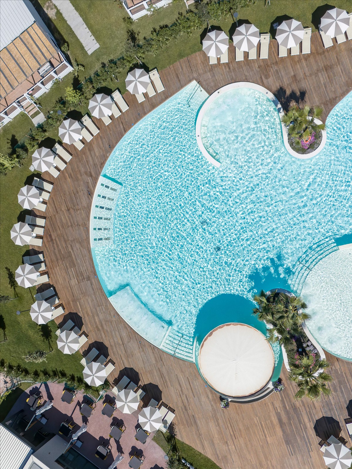 Drone shot of a large, round pool with sun loungers and umbrellas on a wooden deck, surrounded by palm trees and greenery.