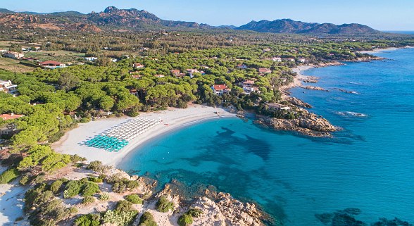 Hotel Sentido Tirreno Resort, Sardinien. An einer schönen Bucht mit weißem Sandstrand, Felsenriffs und kristallklarem Wasser gelegen. Umgeben von Pinienwäldern, im Hintergrund flache Berglandschaft.