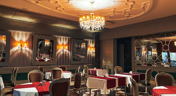 Elegant restaurant with a classic interior, stylish upholstered chairs, red table runners and an opulent chandelier under an artistically designed ceiling.