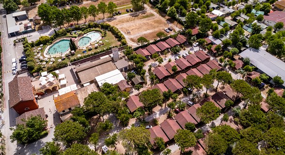 Aerial view of Sentido Tuscany Premium Camp in Toskana, Viareggio with many small wooden huts, lots of greenery and a heart-shaped swimming pool with sun loungers and parasols.
